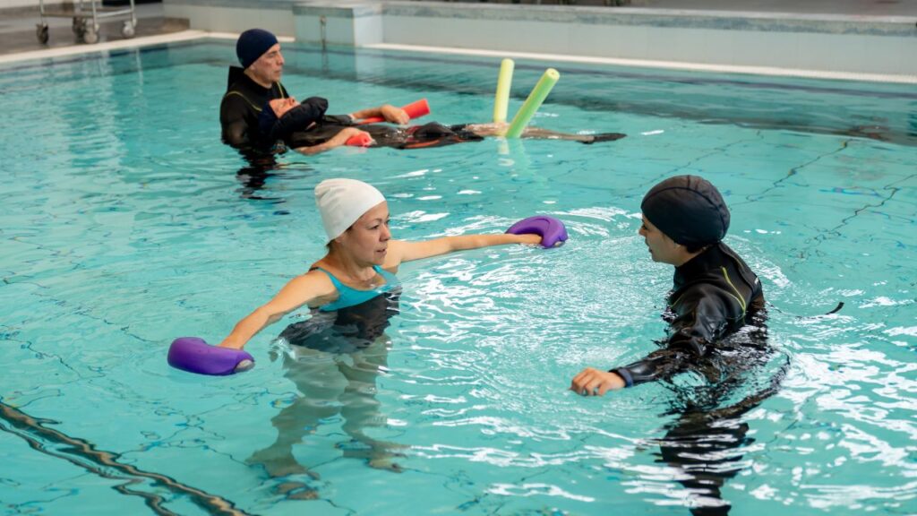 Adults performing guided aqua fitness exercises in a swimming pool using aquatic dumbbells and pool noodles.