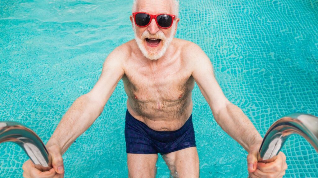 Older man smiling in a swimming pool while holding the pool ladder, representing swimming for healthy aging and confidence in the water.
